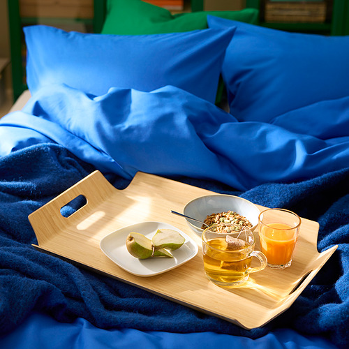 Bandeja de desayuno de bambú en cama azul con fruta, granola, té y jugo.