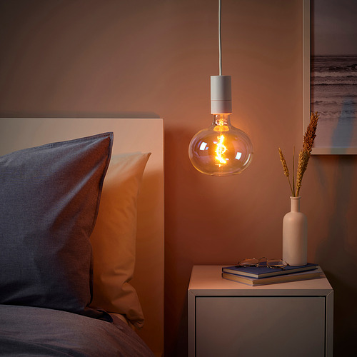 Lámpara SUNNEBY sobre la mesita de noche en una habitación acogedora. Paredes de durazno, almohadas mixtas, cordón limpio, libros, jarrón con hierbas secas. Diseño elegante y minimalista.