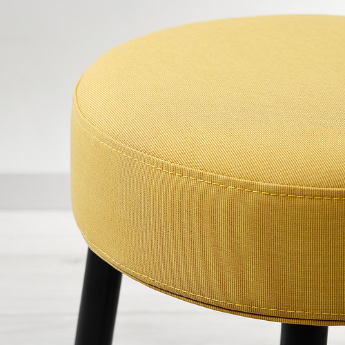Primer plano de un taburete moderno, redondo, tapizado en amarillo con elegantes patas negras, que muestra su tejido de calidad y su diseño limpio.