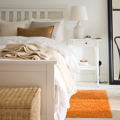 Un dormitorio acogedor con una cama blanca, almohadas neutras y una alfombra naranja de ÄRENDE . Una mesilla de noche tiene una lámpara y objetos, con una silla negra en el borde.
