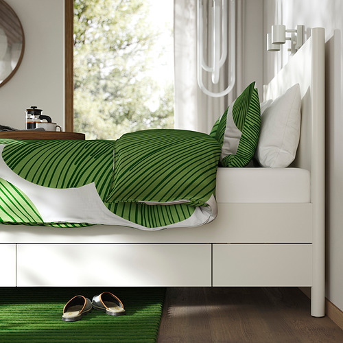Cama con cajones, ropa de cama a rayas verdes, zapatillas, cafetera en mesita de noche, cortinas blancas.