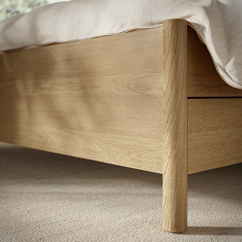Estructura de cama con acabado en madera, con patas redondas y cajón de cierre suave, que se muestra con ropa de cama de color claro.
