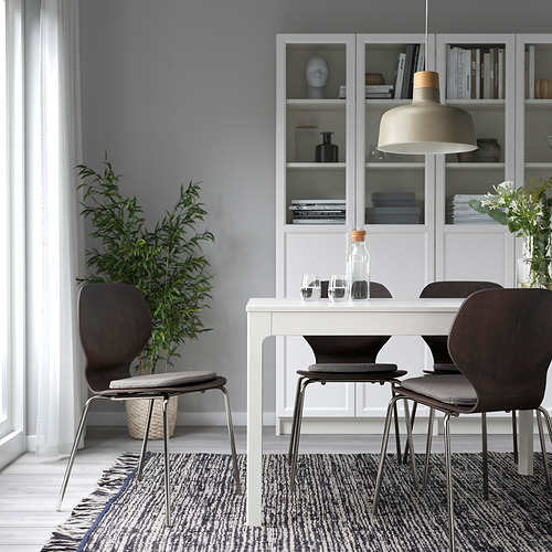Comedor moderno con almohadillas grises, sillas oscuras, mesa blanca, estantería, luz y planta.