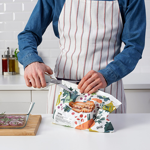 Una persona con camisa vaquera y delantal abre la bolsa con las tijeras MÄRKBART, mostrando su funcionalidad multiusos en la cocina.
