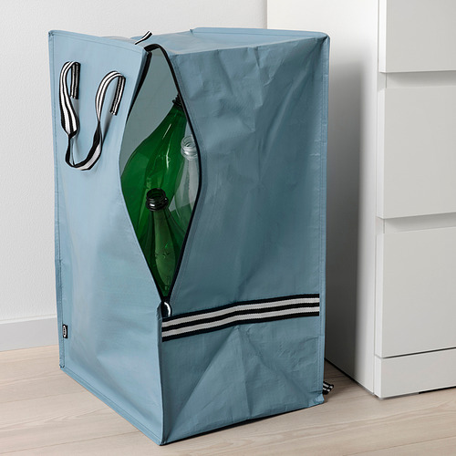 Bolso azul con asas a rayas, cremallera frontal abierta que muestra la botella verde en el interior, se apoya en el armario blanco.