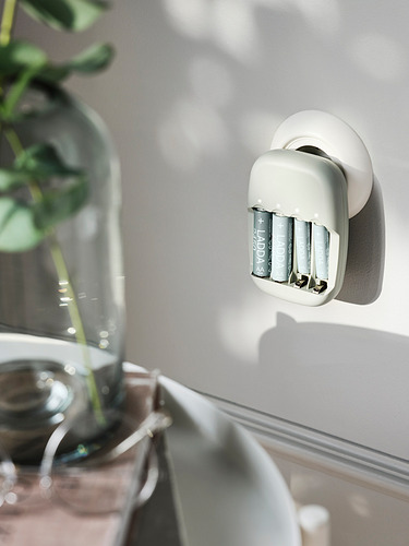 Un cargador de baterías LADDA blanco montado en la pared con tres baterías recargables plateadas en su interior. Parte de una planta verde es visible a su lado.