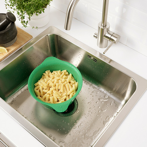 Fregadero de acero inoxidable con colador verde lleno de pasta cocida, colocado en el lado izquierdo. Grifo de plata a la derecha.