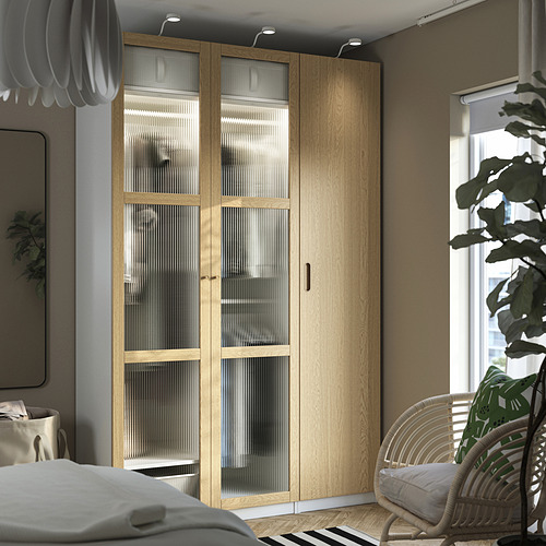 Dormitorio moderno con gran armario PAX beige con puertas de cristal y madera, junto a una silla de mimbre con una planta.