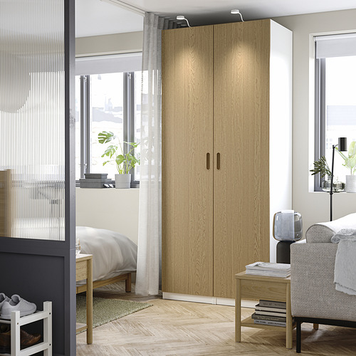 Dormitorio moderno con un gran armario PAX de madera clara, arrinconado entre ventanas, más alto que la cama, con libros y plantas.