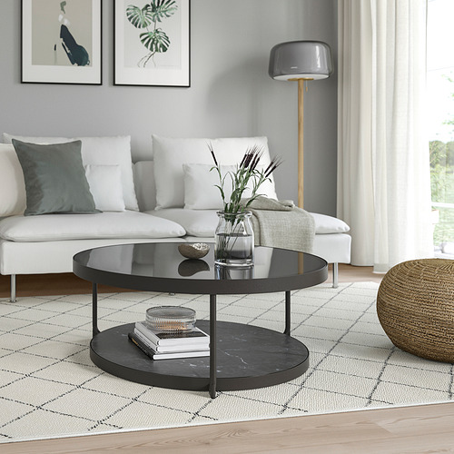 Sala de estar moderna con mesa de centro negra, tapa de vidrio, sofá blanco, lámpara y decoración sobre alfombra estampada.