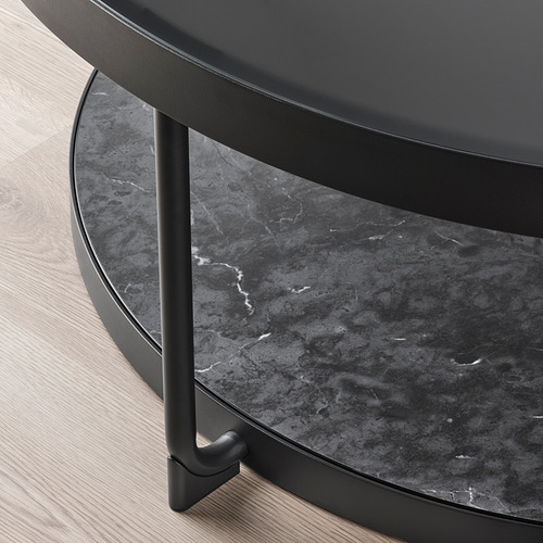 Moderna mesa auxiliar redonda con elegante diseño negro, con tapa de vidrio templado y patas metálicas, se asienta sobre suelo de madera.