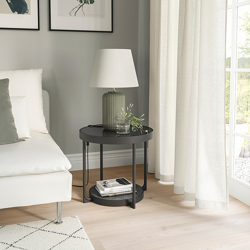 Moderna mesita auxiliar redonda negra con tapa de cristal junto a cama blanca y gran ventanal.
