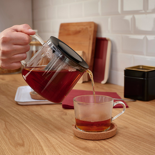 Una mano vierte té de una tetera de vidrio RIKLIG con un infusor en una taza transparente sobre una encimera de madera.