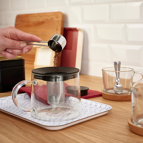 La persona vierte hojas de té en una tetera RIKLIG con infusor en una bandeja.