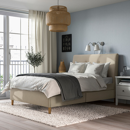 Dormitorio moderno con cama tapizada en beige IDANÄS. Cabecero blando, patas de madera. Ropa de cama gris, alfombra blanca, paredes azul claro. Gran ventanal con cortinas.