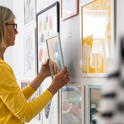Una mujer con camisa amarilla cuelga un marco de fotos colorido en la pared, utilizando numerosas herramientas.