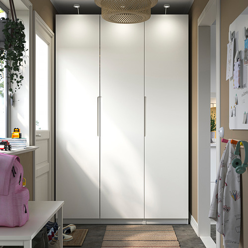 Un gran armario blanco PAX domina la habitación, flanqueado por muebles más pequeños. Las luces brillantes mejoran el diseño limpio. La decoración y los elementos resaltan el espacio organizado.