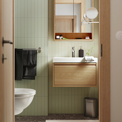 Elegante baño con azulejos verdes, detalles en madera, lavabo, espejo y toalla negra.