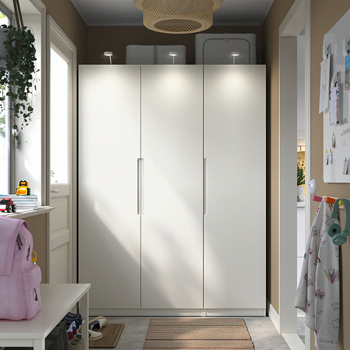 Elegante armario blanco PAX con iluminación integrada, se mantiene alto en la habitación, flanqueado por estantes estrechos y una mochila colorida.
