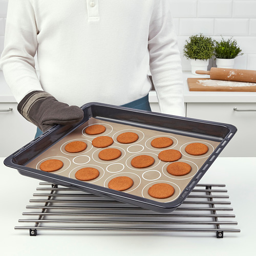 Persona sosteniendo una bandeja para hornear con galletas circulares, colocada en una rejilla para enfriar. Tapete BAKTRADITION visible, que ayuda a hornear de manera uniforme y fácil de liberar.
