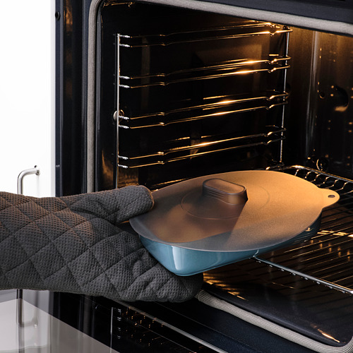 Persona colocando un plato azul con una tapa redonda de silicona marrón en un horno. La tapa ayuda a mantener los alimentos frescos y se puede usar en varios entornos de cocina.