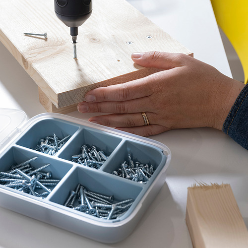 Una mano usa un taladro en una tabla de madera, colocando tornillos de una caja compartimentada que contiene varios tamaños de tornillos.