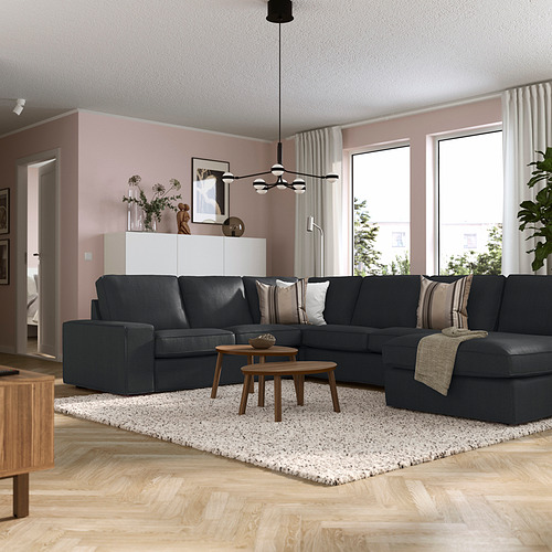 Sala de estar moderna con sofá seccional gris oscuro KIVIK, mesas de madera, paredes rosas, grandes ventanales y cortinas blancas.