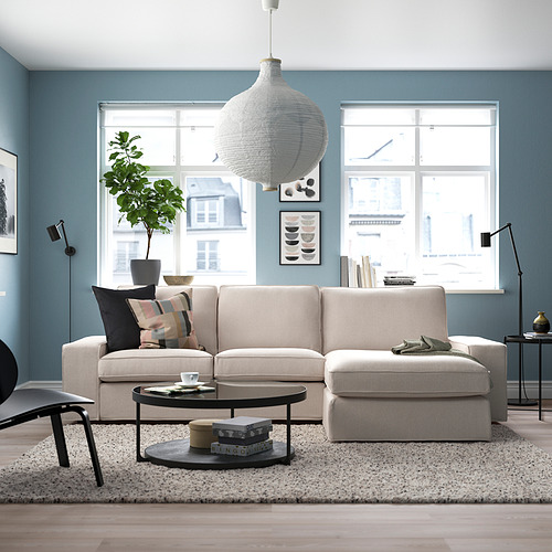 Sala de estar moderna con un gran sofá seccional KIVIK beige, mesa de centro negra, almohadas de colores, alfombra y grandes ventanales.