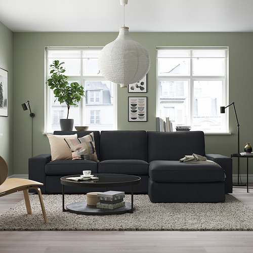 Sala de estar moderna con sofá gris KIVIK, grandes ventanales, plantas y obras de arte abstractas. El sofá es espacioso, con asientos profundos y reposabrazos bajos.