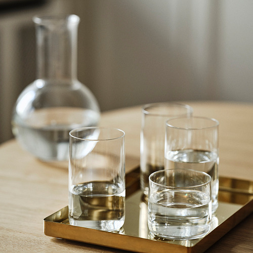 Cuatro vasos elegantes y modernos en una bandeja de oro, llenos de agua. Detrás de ellos, una gran jarra en una cocina minimalista.