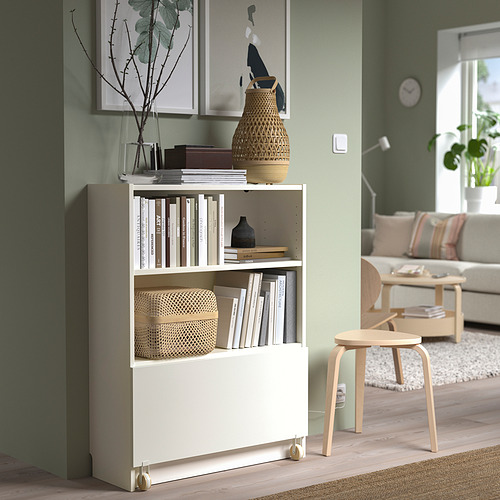 Librería blanca BILLY con ruedas que sostiene libros y jarrones. Más pequeño que el sofá, junto a un taburete de madera.
