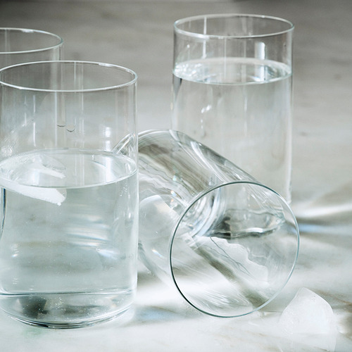 Tres elegantes vasos de vidrio soplado a boca con bordes rectos, cada uno parcialmente lleno de líquido transparente, dispuestos cuidadosamente sobre una superficie lisa.