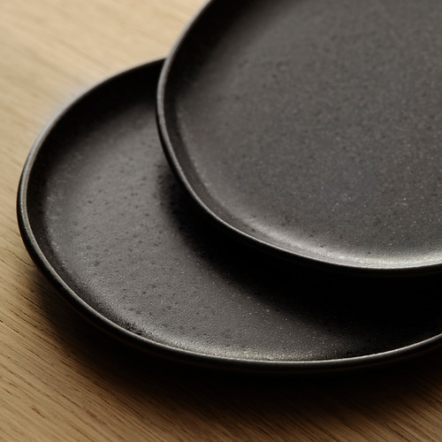 Dos platos negros mate con sutiles motas, apilados ordenadamente sobre una mesa de madera. Aspecto rústico y artesanal.