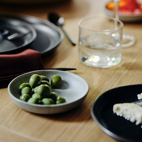 Un pequeño cuenco de guisantes de color verde brillante en un plato gris, un vaso de agua clara y un pequeño plato con comida blanca y desmenuzable junto a ellos sobre una mesa de madera.