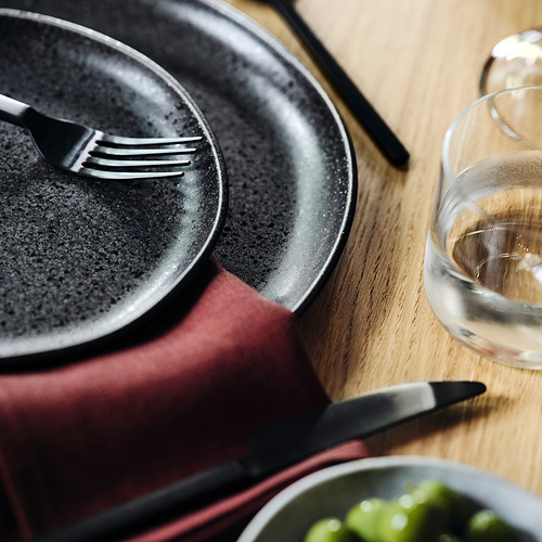 Una mesa con cubiertos, platos y un vaso negros. El tenedor, el cuchillo y la cuchara descansan en los platos; plato verde debajo.