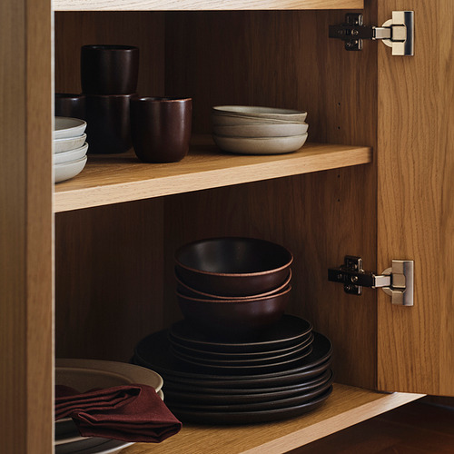 Mueble de madera con platos rústicos marrones; tazones apilados de color marrón oscuro en los estantes medios e inferiores, tazones y tazas más pequeños en el estante superior.