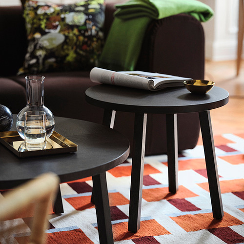 Sala de estar moderna con mesas de café negras, una alfombra tejida a mano con patrón geométrico rojo y blanco, sofá marrón con manta verde y almohadas decorativas.