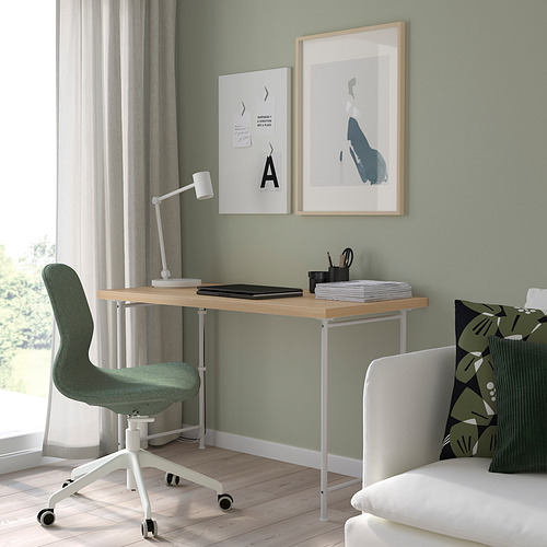 Oficina en casa minimalista con escritorio LAGKAPTEN, silla verde, lámpara blanca, computadora portátil, impresiones artísticas y sofá.
