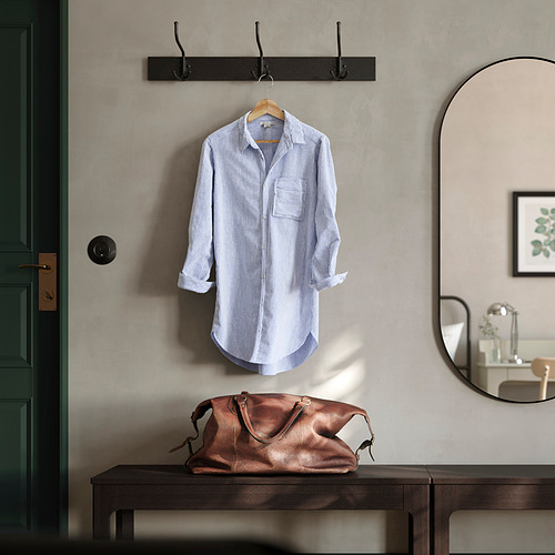 Configuración de la entrada con percha de madera BUMERANG que sostiene una camisa de rayas azules. Bolso de piel marrón oscuro y espejo redondo cerca. Perchero negro arriba.