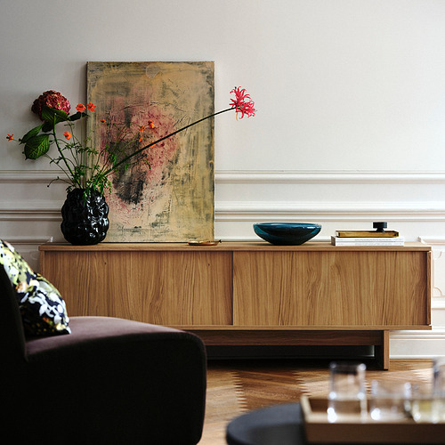 Una sala de estar con muebles de madera, una pintura abstracta y un jarrón de gres negro con flores de colores encima de un armario de madera.