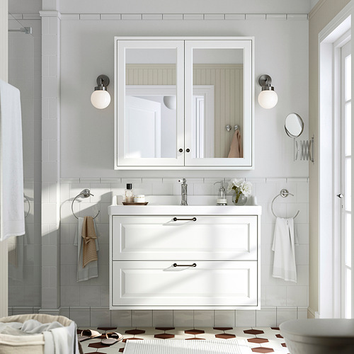 Baño TÄNNFORSEN: tocador blanco, armario con espejo, azulejos estampados. Lavabo flanqueado por apliques, toallas sobre aros de plata, suelo de baldosas geométricas.