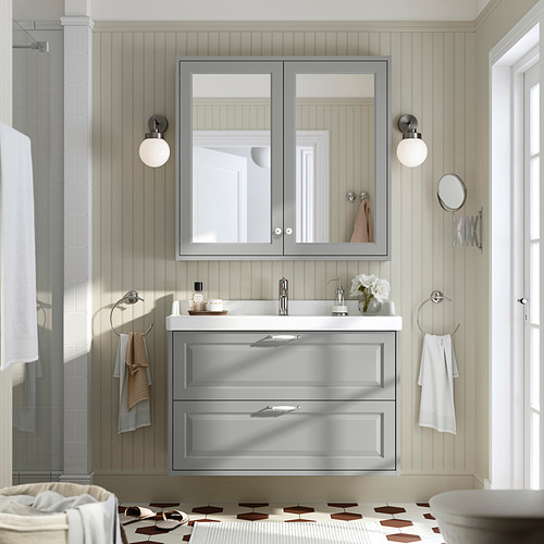 Baño moderno con tocador gris RUTSJÖN, espejo, ducha y lavabo de cerámica. Acentos blancos, apliques y toalleros cromados.