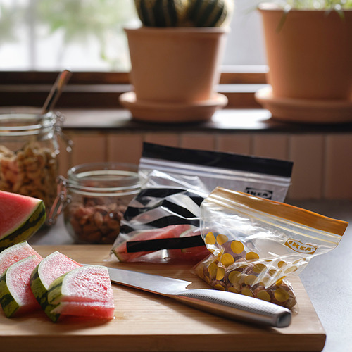 Tabla de cortar de madera con rodajas de sandía, cuchillo, bolsas de comida selladas, frascos y plantas en macetas.