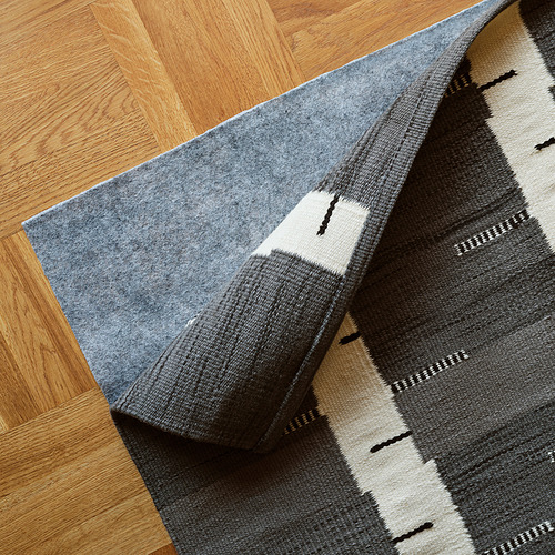 Una alfombra gris tejida a mano con un motivo de abedul descansa sobre un suelo de madera. Parece reversible y de tejido plano para facilitar su uso debajo de los muebles.