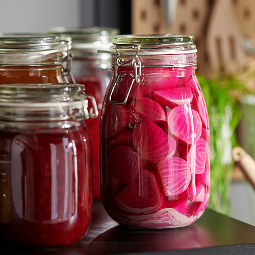 Tres frascos de vidrio con abrazaderas de metal sostienen verduras en escabeche, conservando su frescura. El diseño transparente permite ver fácilmente el contenido.