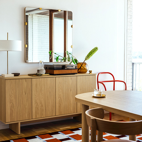 Comedor moderno con aparador de madera, mesa redonda y sillas de colores. Gran espejo sobre el aparador de 2025, sillas rojas y amarillas vibrantes.