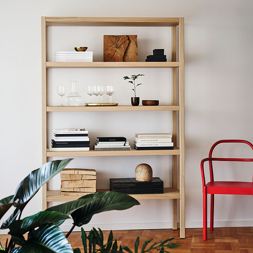 Las estanterías de STOCKHOLM 2025 en madera clara contienen libros, jarrones, plantas; Silla roja alta al lado.