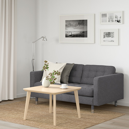 Acogedora sala de estar con sofá gris, mesa de madera, lámpara y fotos. Almohadas blancas y negras, flores, cortinas ligeras y alfombra beige.