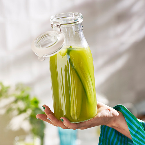 Frasco de vidrio que sostiene en la mano con líquido verde brillante y un pepinillo, sellado con una tapa. El frasco es transparente, muestra el contenido y tiene un sello hermético.