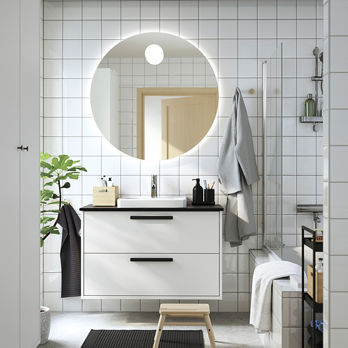 Moderno baño blanco con detalles en negro. Espejo redondo, mostrador flotante, toallas negras, plantas y artículos de tocador.